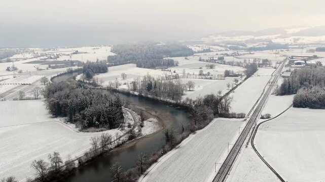 Aerial drone flight over the Reuss River near Sins, Switzerland, showing a snow covered winter landscape with railway line, river and rural countryside.