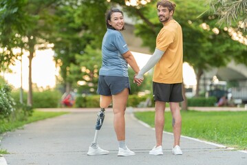 Fototapeta premium Equality and diverse friendship disabled woman with prosthetic leg walking with supportive male in public park. Helping man holding hands and guiding female amputee friend during outdoors .