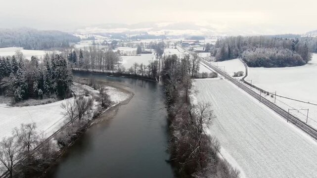Aerial drone flight over the Reuss River near Sins, Switzerland, showing a snow covered winter landscape with railway line, river and rural countryside.