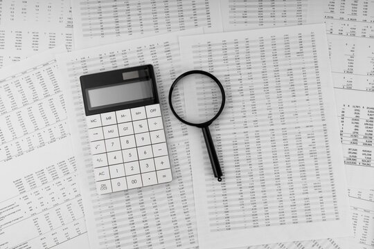 Calculator and magnifying glass on top of a stack of complex financial data sheets with calculator, magnifying