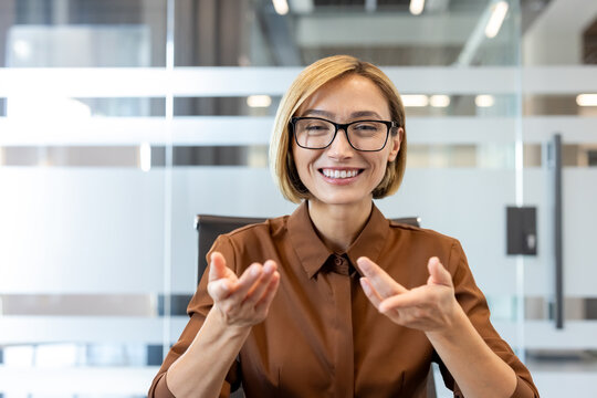 Woman wearing glasses and a brown shirt talking during a virtual meeting, communicating effectively in a modern office environment
