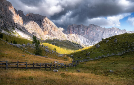 Wooden fence on green meadow with dirt road leading to mountain peaks in Dolomites, Italy. Alpine summer landscape under stormy dark clouds with dramatic sunlight hitting rocky cliffs and valley