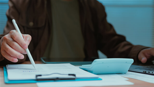 Person holding stylus reviewing financial document near calculator calculating budget task workplace interior employee analyzing data corporate report startup strategy project management commercial.