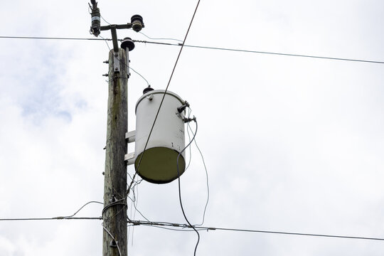 Electric distribution transformer attached to wooden pole with power line connections against cloudy sky background. Power energy supply concept.