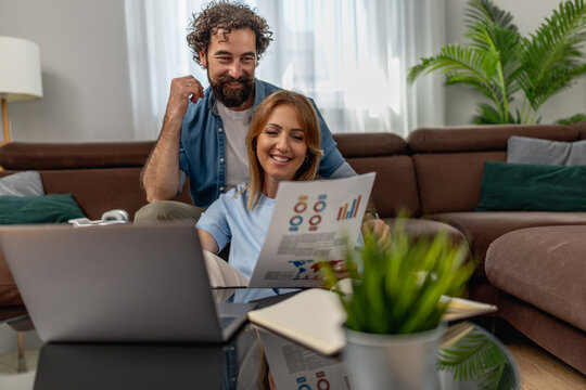 Couple works together at home while looking at documents on a laptop in a living room