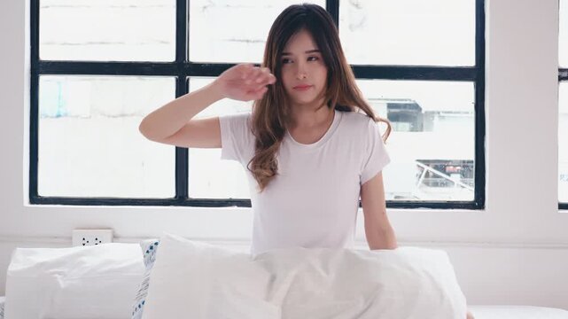 Cute young Asian woman waking up in bed, yawning and rubbing her eyes, then smiling and waving hand saying hello to camera. Happy female in white t-shirt enjoying morning routine