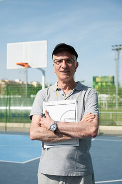 Senior male coach smiling, posing with clipboard on an outdoor basketball court