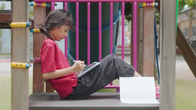 Asian kid enjoying a relaxing hobby, drawing digital art on tablet at outdoor school playground. Selective focus highlights creative elementary student spending personal leisure time with technology.