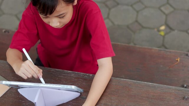 Top view of Asian kid using stylus pen on a digital tablet for e-learning at outdoor school table. It shows the young student engaging with technology for modern education and digital study concepts.