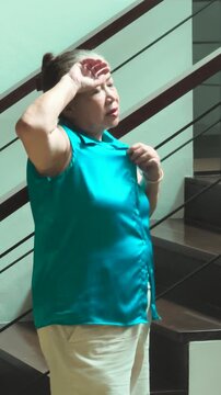 Tired and dizzy elderly asian woman wiped out by summer heat inside her home, fan herself with her shirt to cool down, vertical video