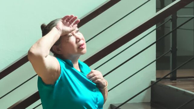 Tired and dizzy elderly asian woman wiped out by summer heat inside her home, fan herself with her shirt to cool down