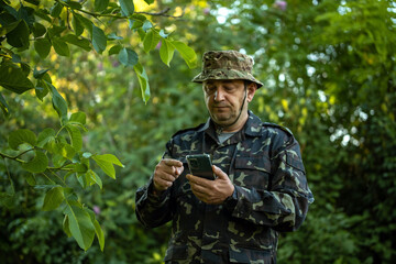 Fototapeta premium A soldier in a camouflage uniform stands in a forested area. He is looking at a smartphone while surrounded by trees and plants during daylight