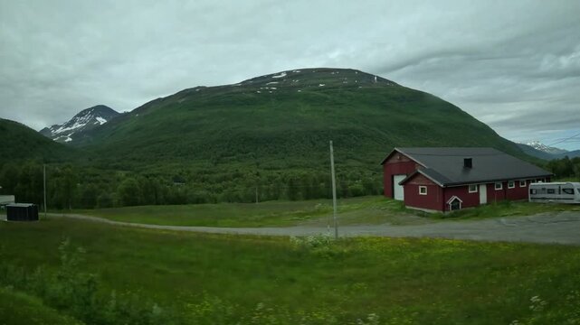 Driving side window view of traditional red barn with caravan and green mountains in Northern Norway