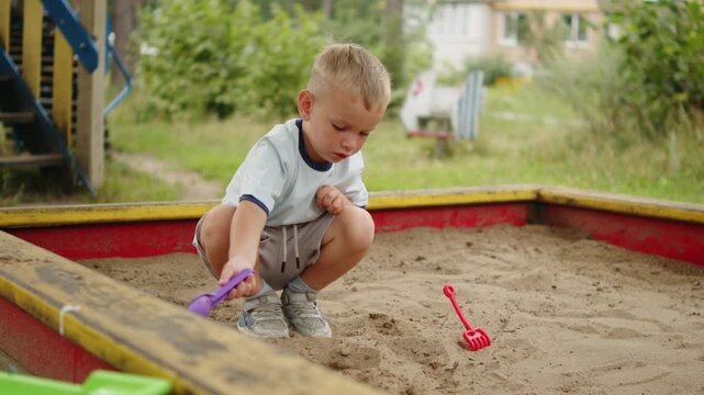 Little boy playing in a sandbox. Small blonde child in a wooden sandbox, digging in the sand with a purple plastic toy shovel. Cute kid having fun on a playground, child entertained in a sandpit.