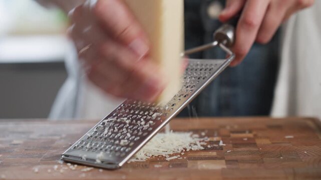 Woman grates block of hard cheese with metal grater over wooden cutting board. Food preparation for a homemade meal
