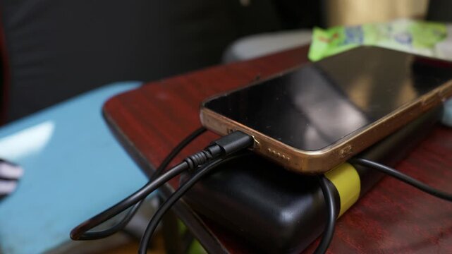 A smartphone charges from a portable power bank on a table at an outdoor campsite with a fly sitting on the cable. Concept of off-grid energy, portable technology, and outdoor leisure.