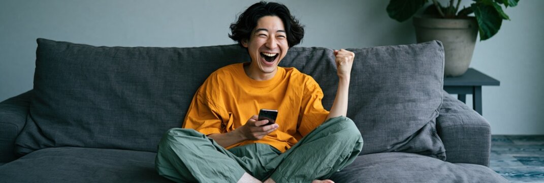 Young man laughing and cheering with mobile phone on cozy couch for banner and website background use