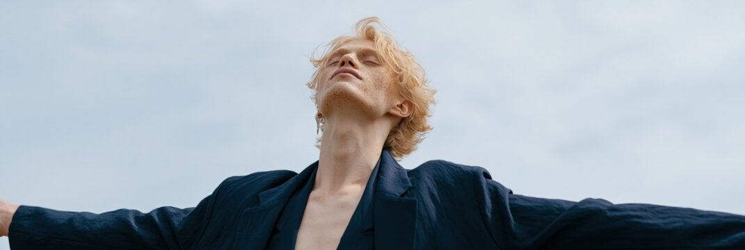 Blonde young man with open jacket and raised arms against pale sky background for editorial banner and fashion header