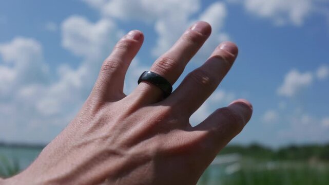 A man hand with a dark ring reaches toward a bright blue sky filled with white clouds. Soft sunlight illuminates the skin and highlights the veins on the back of the hand in nature.