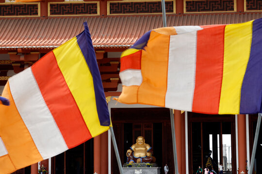 Quoc An Khai Tuong Pagoda. Colorful Buddhist flags flutter in front of a temple, symbolizing peace and spirituality. A golden statue of the Laughing Buddha can be seen inside. Long Phuoc, Vietnam.