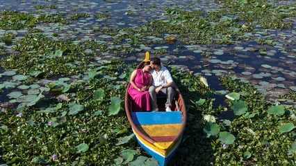 Fotobehang Lotusbloem Aerial drone view of a couple enjoying a colorful boat ride across a scenic lake covered with lotus leaves and aquatic plants, surrounded by lush greenery.  © Vishal