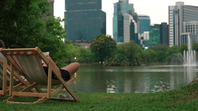 Person reading and relaxing in deck chair at park lake with fountain, Peaceful pond leisure with modern city skyline in Lumpini Park, Bangkok Thailand 4k
