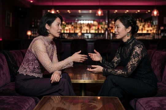 Two elegant women conversing in a dimly lit lounge bar with purple ambient lighting and blurred bar background
