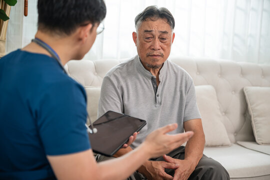 Home Healthcare Consultation for an Asian Senior Man Seated on a Sofa while a Male Caregiver in Blue Scrubs Discusses His Condition and Uses a Digital Tablet