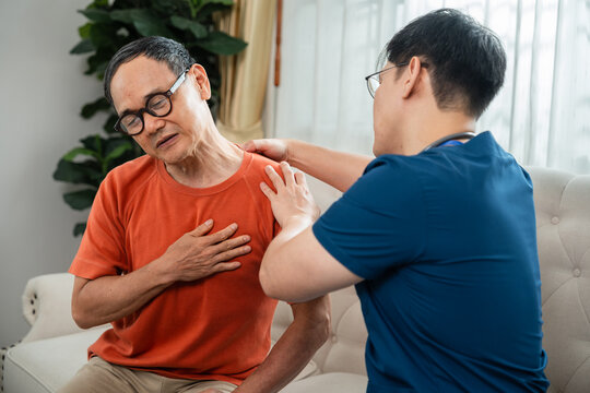 Senior Care for an Asian Elderly Man Holding His Chest with a Distressed Expression while a Male Caregiver in Blue Medical Scrubs Provides Support at Home