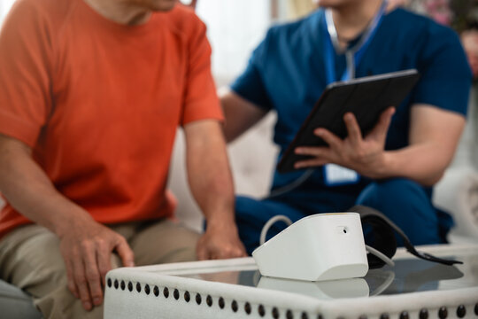 Home Healthcare Consultation for an Asian Senior Man as a Male Caregiver in Blue Medical Scrubs Holds a Digital Tablet beside an Electronic Medical Device at Home