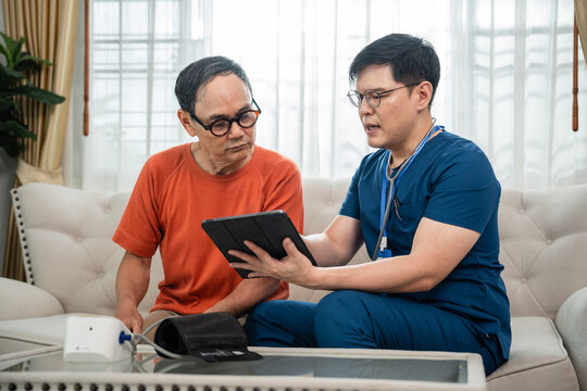 Home Healthcare Consultation for an Asian Senior Man as a Male Caregiver in Blue Medical Scrubs Reviews Information with Him on a Digital Tablet at Home