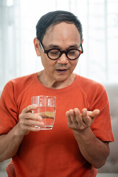 Senior Medication Routine of an Asian Elderly Man Holding a Glass of Water and Looking at Tablets in His Hand while Seated in a Bright Home Interior