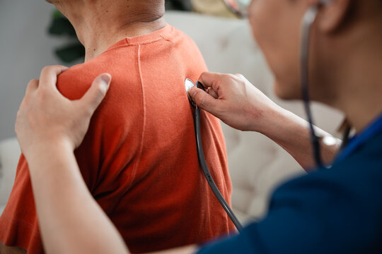 Home Healthcare Examination for an Asian Senior Man as a Male Caregiver in Blue Medical Scrubs Uses a Stethoscope to Check His Upper Back and Shoulder at Home