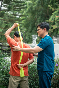 Senior Physical Therapy for an Asian Elderly Man Performing Resistance Band Shoulder Exercises while a Male Caregiver in Blue Scrubs Guides Him in an Outdoor Garden