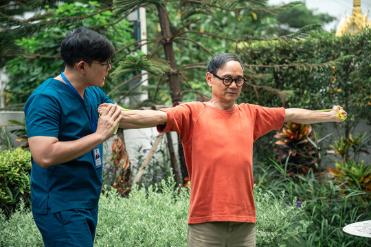Senior Physical Therapy for an Asian Elderly Man Performing Resistance Band Shoulder Exercises while a Male Caregiver in Blue Scrubs Guides Him in an Outdoor Garden