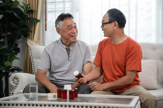 Senior Medication Management with Two Asian Elderly Men Discussing a Medicine Bottle while Seated Together beside Additional Medication in a Bright Home Interior