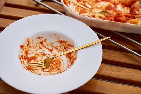 Empty plate with tomato sauce remains and fork beside baking dish with leftover gnocchi