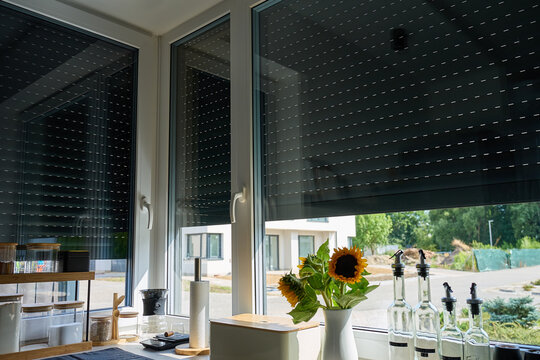 Roller shutters are partly lowered over kitchen windows beside sunflowers, jars and glass bottles