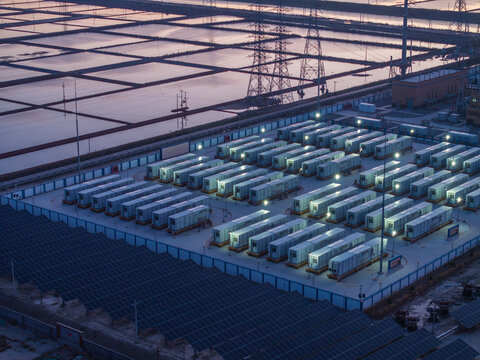 Large-scale energy storage station and photovoltaic array at dusk with illuminated modules and surrounding industrial infrastructure
