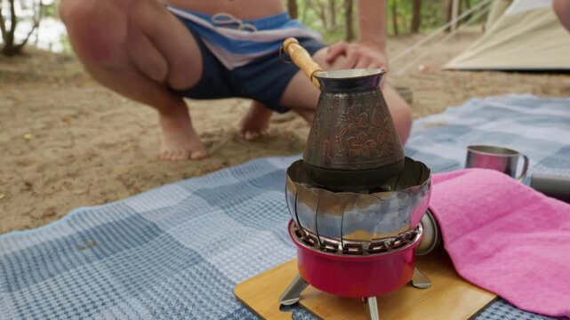 A traditional copper coffee pot heats up on a small portable gas stove placed on a picnic blanket outdoors. In the background, a camper relaxes near a tent during a morning break.