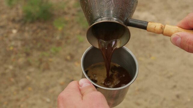 Fresh Turkish coffee pours smoothly from a traditional cezve into a metal camping mug outdoors. Hands hold the metal cup and wooden handle while enjoying a warm morning beverage in nature.