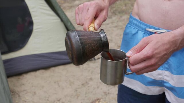 A man pours freshly brewed Turkish coffee from a cezve into a metal mug at a campsite. Concept of morning caffeine ritual, outdoor camping life, leisure, and adventure lifestyle.
