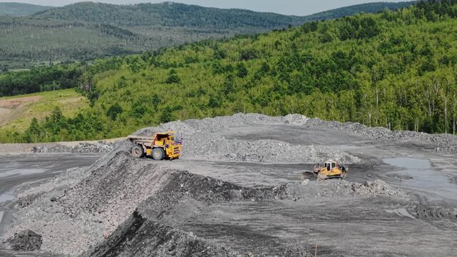 Aerial tracking shot of dump truck unloading copper, gold ore at mountain quarry pit with loader nearby panoramic pan over industrial excavation site.
