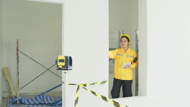 Renovation site worker in yellow jacket measuring room interior wall opening unfinished room repair work with laser level safety tape and inspection notes