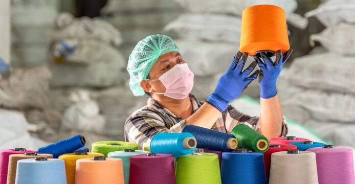 Textile factory male worker in mask hairnet and gloves handling and checking colorful yarn spool for quality control Industrial textile inspection with focus in warehouse