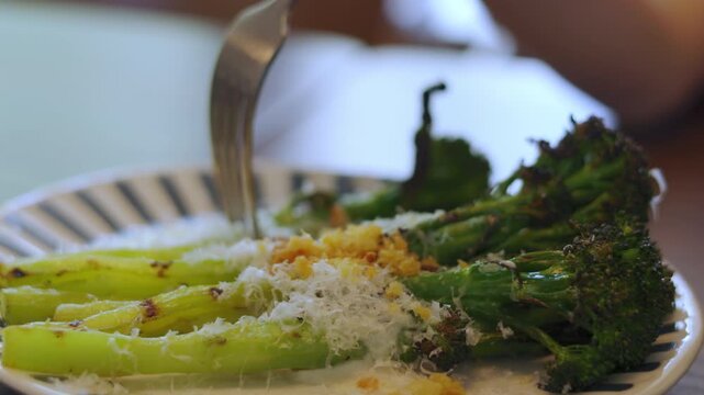 Close up of a fork piercing delicious grilled broccolini topped with parmesan cheese. Healthy roasted green vegetable dish served on a plate. Shot with selective focus. Gourmet vegan dining concept.