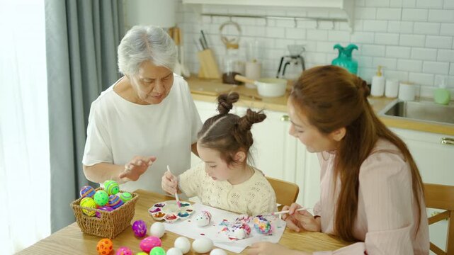 Happy multi-generational Asian family painting colorful Easter eggs together in a bright modern kitchen. Grandmother, mother, and a cute little girl bonding over fun holiday craft activities.