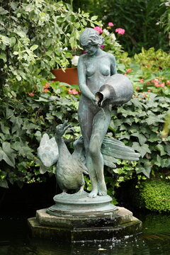 Bronze garden fountain statue of a woman pouring water from a jug beside a goose in a lush ornamental garden with flowing water.