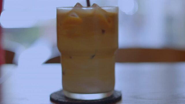 Close up of a straw stirring refreshing iced coffee in a glass. Cold milk latte with ice cubes in natural light. Shot with selective focus. Morning cafe beverage and summer cooling drink concept.