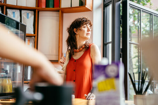 Curious middle-aged woman in orange dress waiting at sunny specialty coffee shop